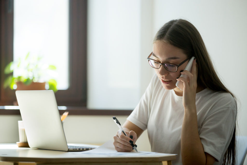 Busy female worker talking over phone, consulting client, solving company problems, giving help and assisting while writing down important information. Call center operator
